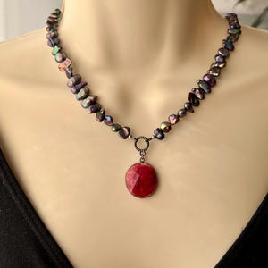 May include: A necklace featuring iridescent, dark-colored freshwater pearls. The necklace has a silver clasp and a red, round pendant. The pendant is set in a silver frame. The necklace is displayed on a mannequin.