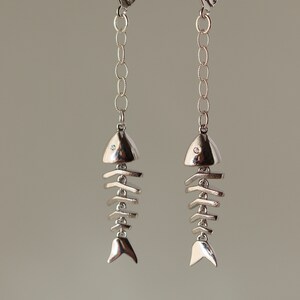 Op de afbeelding: Zilveren visgraat oorhangers met een kleine, heldere steen op elke vis. De oorhangers hangen aan een zilveren ketting en hebben een sluiting met een hendel.