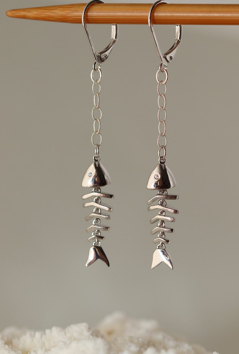 Op de afbeelding: Zilveren visgraat oorhangers met een kleine, heldere steen op elke vis. De oorhangers hangen aan een zilveren ketting en hebben een sluiting met een hendel.