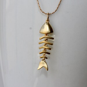 Puede incluir: Un collar de espinas de pescado dorado con un peque&ntilde;o acento de diamante en la cabeza del pez.