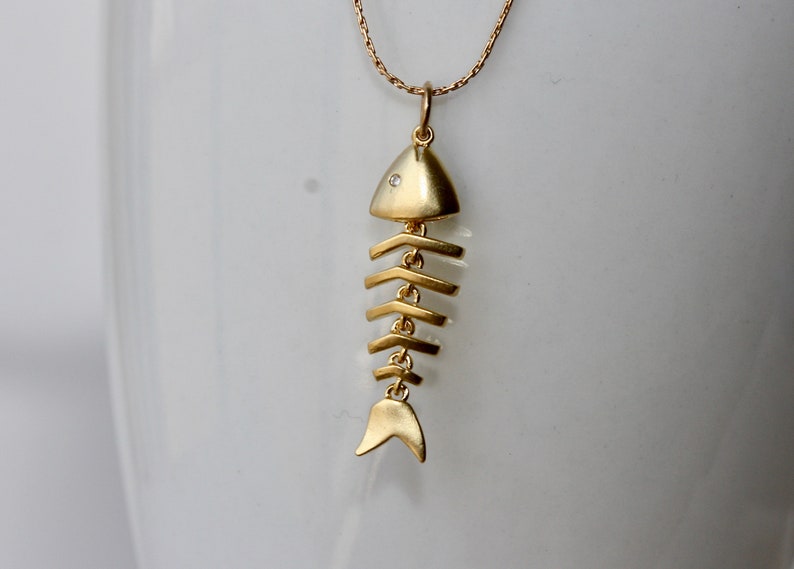 Puede incluir: Un collar de espinas de pescado dorado con un peque&ntilde;o acento de diamante en la cabeza del pez.