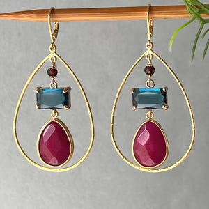 May include: A pair of gold-toned teardrop earrings featuring a central rectangular blue gemstone, a small dark red bead above it, and a larger faceted teardrop-shaped magenta gemstone suspended within the open gold frame.