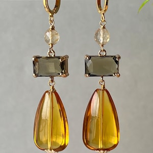 May include: A pair of gold-tone hoop earrings featuring a faceted clear bead, a rectangular dark gray stone, and a large amber-colored teardrop-shaped pendant. The components are connected by gold-tone wire and metal links in a vertical arrangement.