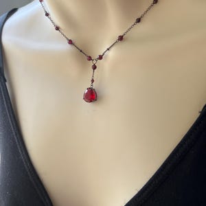 May include: A delicate necklace with a single large red gemstone pendant. The necklace is made of black chain with small red beads.