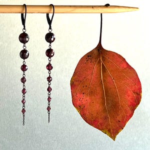 May include: A pair of dangle earrings with a long chain of dark red beads and black metal accents. The earrings are hanging from a wooden dowel rod. A dried, red leaf is hanging from the dowel rod.