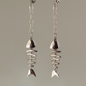 Op de afbeelding: Zilveren visgraat oorhangers met een klein, helder steentje bovenop elke vis. De oorhangers hangen aan een zilveren ketting.