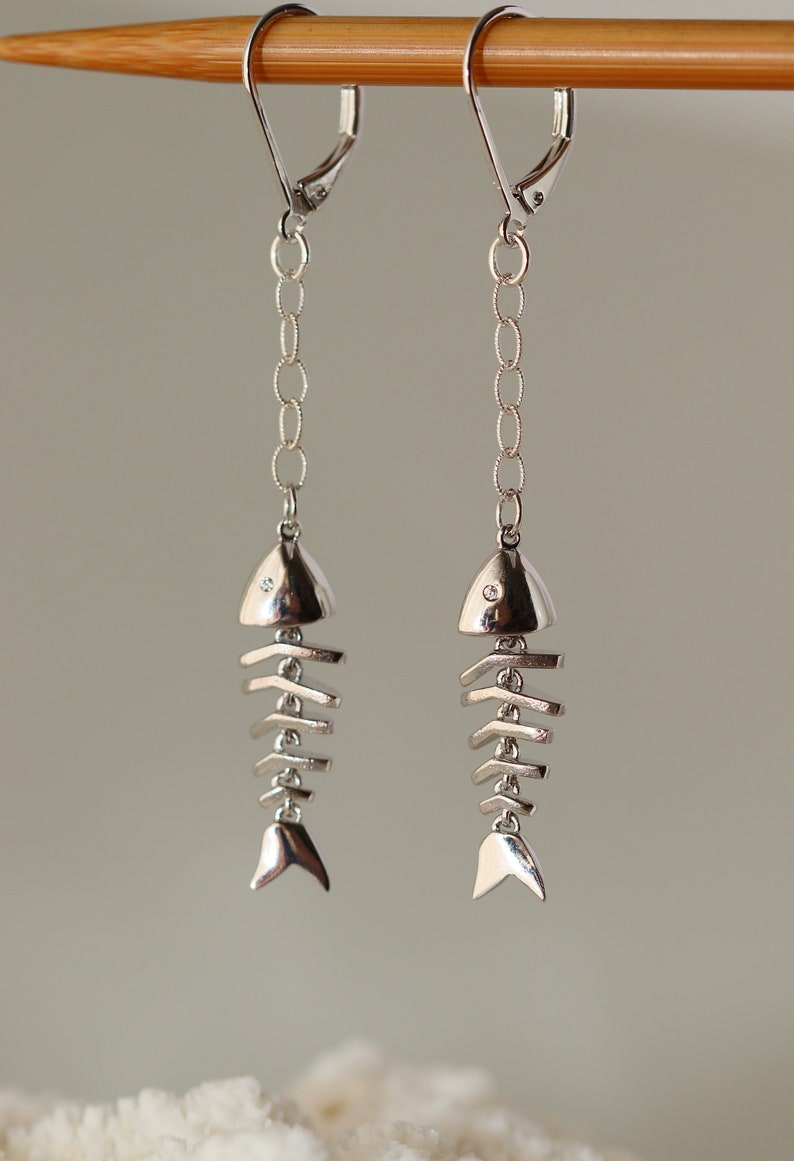 Op de afbeelding: Zilveren visgraat oorhangers met een klein, helder steentje bovenop elke vis. De oorhangers hangen aan een zilveren ketting.
