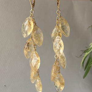 May include: A pair of dangle earrings with gold-colored hooks and chains. Each earring features multiple faceted, teardrop-shaped, light yellow gemstones. The earrings are displayed against a neutral gray background.