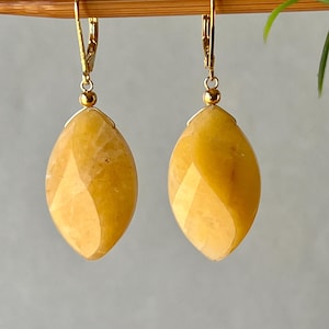 May include: A pair of drop earrings featuring faceted, marquise-shaped yellow gemstones suspended from gold-toned leverback hooks. Each stone is capped with a small gold bead, showcasing a translucent, warm golden hue with natural internal variations.