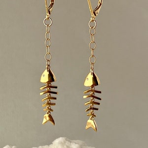 Op de afbeelding: Gouden visgraat oorhangers met een klein diamant accent op elke vis. De oorhangers liggen op een houten oppervlak.