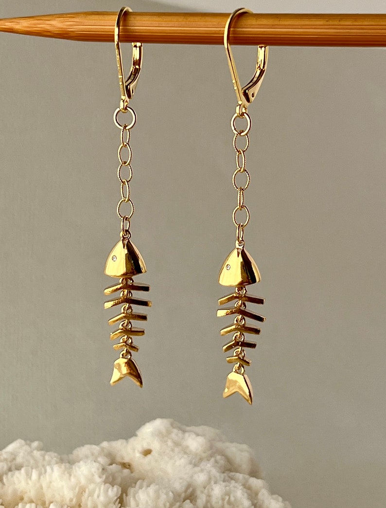 Op de afbeelding: Gouden visgraat oorhangers met een klein diamant accent op elke vis. De oorhangers liggen op een houten oppervlak.