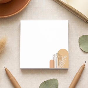 May include: A square white sticky note pad featuring a graphic design in the bottom right corner consisting of three tan and beige arches with thin brown line details and a stylised botanical illustration of dried grass fronds.