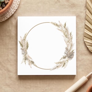 May include: A square white card featuring a central gold circular frame adorned with a wreath of dried pampas grass and palm leaves. The card is positioned alongside a succulent in a terracotta pot, a dried palm fan, and a wooden pen.