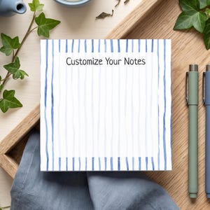 Könnte beinhalten: Ein quadratischer Notizblock mit weißer Mitte und einem blauen vertikal gestreiften Rand. Der Text 'Customize Your Notes' ist oben mittig in Schwarz aufgedruckt. Der Notizblock liegt auf einem Holztablett neben drei Stiften in Salbeigrün, Blau und Schwarz.