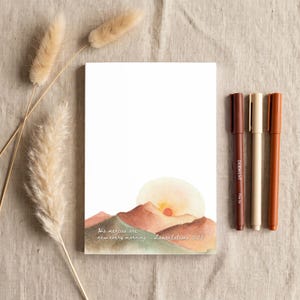 May include: A rectangular notepad featuring a watercolor illustration of rolling mountains and a sun at the bottom. The text reads, "His mercies are new every morning Lamentations 3:23." Three pens in shades of brown and cream are positioned to the right.