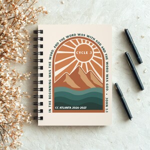 May include: A spiral-bound notebook with a cream cover featuring a graphic of a sun, mountains, and waves in shades of orange, brown, and teal. The text reads: IN THE BEGINNING WAS THE WORD, AND THE WORD WAS WITH GOD AND THE WORD WAS GOD. JOHN 1:1, CYCLE 3, CC ATLANTA 2026-2027.