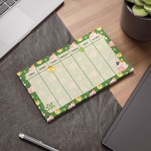 May include: A rectangular weekly planner notepad with a green border featuring illustrations of cats, potted plants, and mugs. The layout includes columns labeled Sunday through Saturday, with a light cream background and a repeating pattern of small icons.