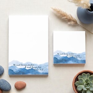 May include: A rectangular notepad and a square notepad, both featuring a white background with a blue watercolor mountain landscape at the bottom. The text "He must become greater; I must become less. John 3:30" is printed in dark blue script.