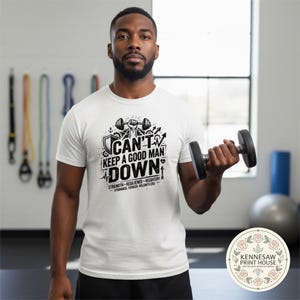 May include: A white t-shirt with the text "CAN'T KEEP A GOOD MAN DOWN" in black. The shirt features a barbell graphic and the words "STRENGTH - RESILIENCE - RECOVERY". The person in the image is holding a black dumbbell.