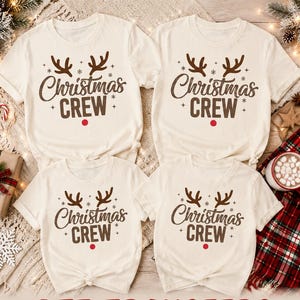 May include: Four cream-colored short-sleeve t-shirts featuring a brown graphic print of reindeer antlers, the text "Christmas CREW," and a small red dot. The shirts are arranged in a group, displaying a consistent holiday-themed design.