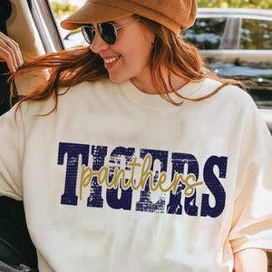 May include: A cream-colored crewneck sweatshirt featuring the word TIGERS in large, distressed navy blue block letters with the word panthers written in a gold cursive script layered over the center. The garment has a relaxed fit.