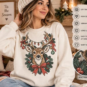 May include: A cream-colored sweatshirt featuring a graphic of a deer head surrounded by a wreath of pine branches, red berries, and a large red bow. The deer antlers are decorated with string lights and ornaments. Text includes DTF TRANSFER.