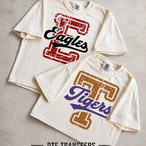 May include: Two off-white short-sleeve t-shirts. The top shirt features a distressed red letter E with the black script text Eagles. The bottom shirt features a distressed brown letter T with the purple script text Tigers. Text reads DTF TRANSFERS.