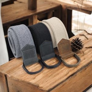 May include: Four fabric camera straps in gray, black, and beige, with leather and metal hardware. The straps are rolled up and displayed on a wooden surface, showcasing their different colors and textures. The leather pieces are in gray, black and brown.