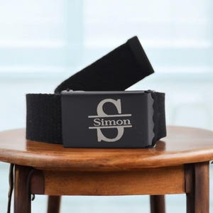 May include: A black canvas belt with a silver buckle engraved with the letter "S" and the name "Simon".