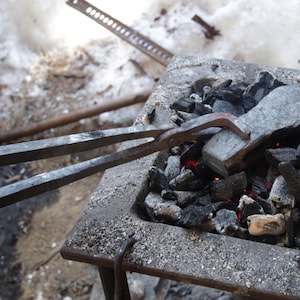 Könnte beinhalten: Ein Metallobjekt wird in einer Schmiede erhitzt. Das Objekt wird mit einer Zange gehalten. Die Schmiede ist mit glühenden Kohlen gefüllt und es steigt Rauch aus der Schmiede auf.