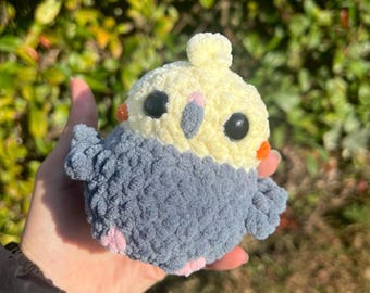 May include: A handmade crochet bird in the shape of a cockatiel. The bird has a yellow head with black eyes, orange cheeks, and a gray and blue body. The bird is made of soft yarn and has a fluffy texture.
