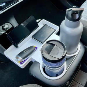 Könnte beinhalten: Ein Auto-Konsolen-Organizer mit einem weißen Tablett, das ein schwarzes Smartphone, eine Brieftasche und einen Autoschlüssel hält. Eine graue Wasserflasche und ein schwarz-blauer Reisebecher befinden sich in den Getränkehaltern. Der Organizer befindet sich in einem Autoinnenraum.