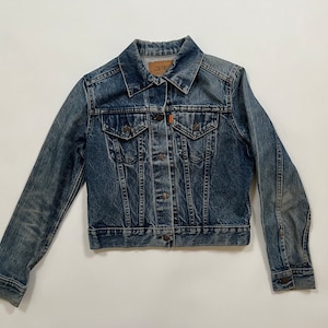Op de afbeelding: Een blauw denim jack met knoopsluiting, twee borstzakken en twee voorzakken. Het jack heeft een verbleekte look en is in goede staat.