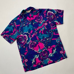 Puede incluir: Una camisa de manga corta con un estampado floral vibrante en tonos de rosa, azul y morado. La camisa tiene un cierre de botones en la parte delantera y un cuello.