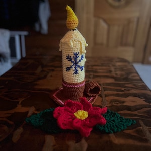 May include: Crocheted candle with a yellow flame, cream-colored wax, and a blue snowflake design. The candle sits in a brown holder with a red and yellow poinsettia and green leaves at the base. The candle is a decorative item.