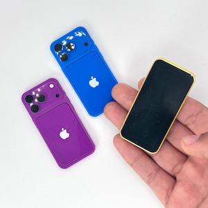 Puede incluir: Tres fundas de teléfono en miniatura en azul y morado, y un pequeño teléfono negro con un borde dorado. Las fundas tienen un diseño de protuberancia de cámara y un logotipo de Apple blanco. El teléfono negro se sostiene en una mano.