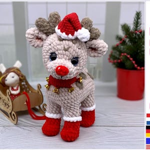 May include: Crocheted reindeer toy wearing a red and white Santa hat with jingle bells around its neck. The reindeer is standing on a wooden surface.