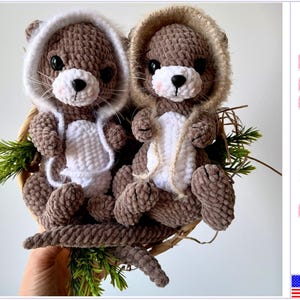 Può includere: Due giocattoli amigurumi di lontra all'uncinetto con pelliccia marrone e cappucci bianchi. Le lontre sono sedute in un cesto intrecciato con fogliame verde.