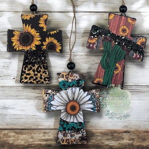 May include: Three cross-shaped hanging ornaments featuring distinct patterns. One displays sunflowers and leopard print, another shows a cactus with a black glitter band, and the third features a large white daisy with turquoise and leopard print accents.
