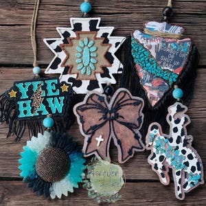 May include: A collection of decorative air fresheners with a Western theme. Designs include a steer, a bow, a sunflower, and shapes with cow print, turquoise accents, and the text "Yee Haw" and "Cowboy Up or Shut Up Baby."
