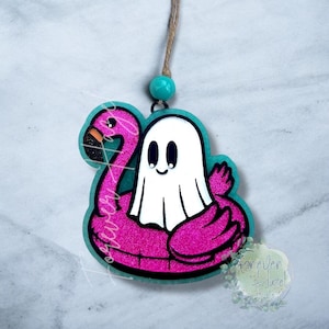 May include: A hanging ornament featuring a white ghost sitting inside a bright pink glittery flamingo pool float. The design is outlined in teal and includes a teal bead attached to a brown twine string for hanging. The graphic has a smooth finish.