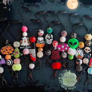 May include: A collection of Halloween-themed beaded lanyards with various charms, including pumpkins, skulls, and cartoon characters. Each lanyard features a black cord and a metal clasp. Some charms have the text "BULL SHEET" and "VIBING WITH MY DEMONS".