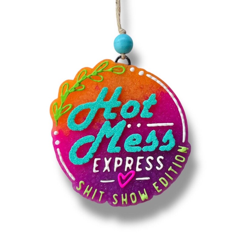 Puede incluir: Un adorno colgante redondo con un fondo degradado brillante en naranja, rosa y morado. Presenta el texto turquesa "Hot Mess EXPRESS" y "SHIT SHOW EDITION" en blanco, con un peque&ntilde;o coraz&oacute;n rosa y detalles de hojas verdes, suspendido por un cordel con una cuenta azul.