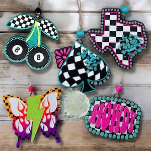 May include: Five hanging ornaments featuring a cherry shape with 8-ball designs, a spade shape with turquoise accents, a Texas state shape with turquoise, a butterfly with a lightning bolt, and a rectangular sign with the word HOWDY in pink.