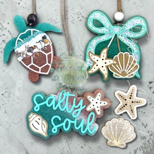May include: Three hanging ornaments and two loose pieces featuring coastal themes. One ornament is a brown sea turtle with a white shell pattern and sand, another is a teal glitter bow with a starfish and shell, and a sign reads salty soul.