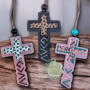 May include: Three hanging cross ornaments with twine loops and beads. The left cross is light pink with a blue leopard print and glitter. The center cross is dark gray with a brown leopard print. The right cross features pink and teal glitter.
