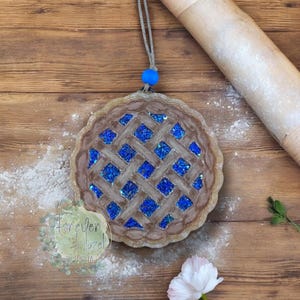 May include: A round, brown, pie-shaped ornament featuring a lattice crust design filled with sparkling blue glitter. The ornament hangs from a thin, light-colored string with a single bright blue bead attached at the top for decorative hanging.