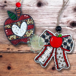 May include: Two hanging ornaments: one apple-shaped with red glitter, leopard print stripes, and a central white heart; the other a bow-shaped ornament featuring a red apple, black and white checkered pattern, leopard print, and the word teacher.