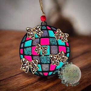 May include: A round hanging ornament featuring a grid pattern of hot pink, teal, and silver glitter squares. Four leopard print bows are attached to the front. The ornament hangs from a jute string with a single red bead at the top.
