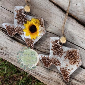 May include: Two Texas-shaped ornaments made of clear resin filled with copper-colored glitter. One ornament features a large yellow sunflower with a dark brown center. Each piece includes a natural wooden bead and a jute twine loop for hanging.